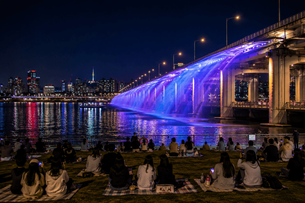 Banpo Bridge Rainbow Fountain at night with people watching from Han River park