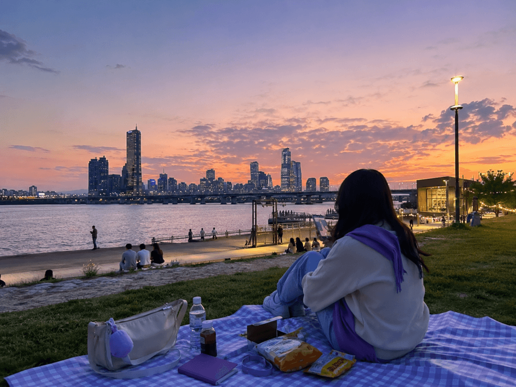 Han River ARMY Route ARMY traveler relaxing by the Han River in Seoul at sunset with a picnic mat, skyline and soft purple travel accents