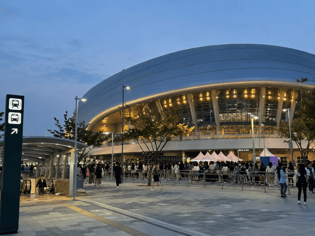 Large concert arena in Seoul at evening with fans waiting outside and public transportation signs nearby