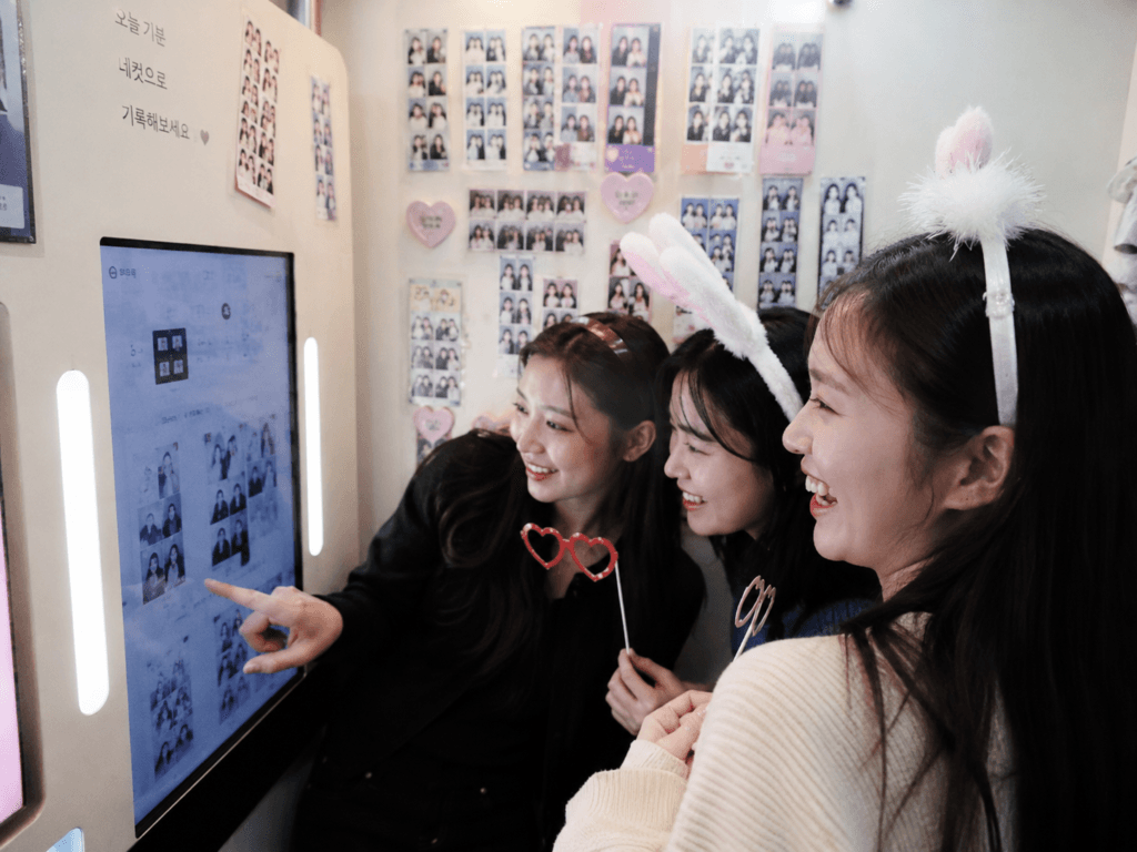 Friends choosing poses and frames inside a Korean photo booth with cute props, sample photo strips and bright lights