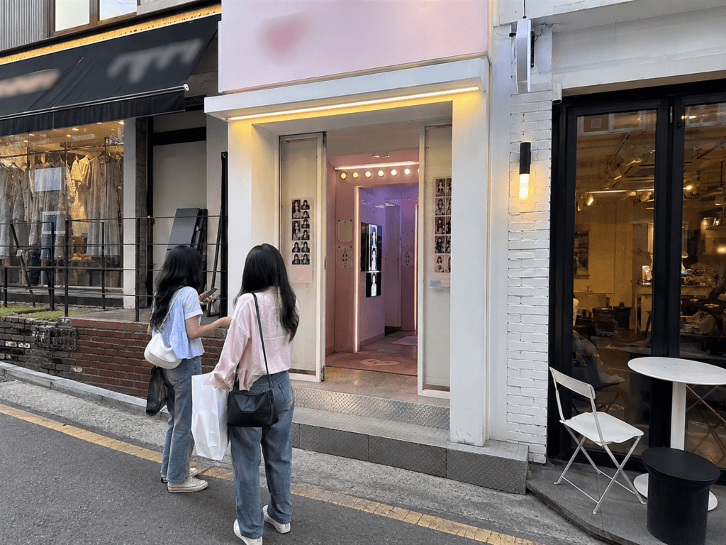 Small Korean photo booth shop on a trendy Seoul street with two travelers standing outside and nearby cafes and fashion stores
