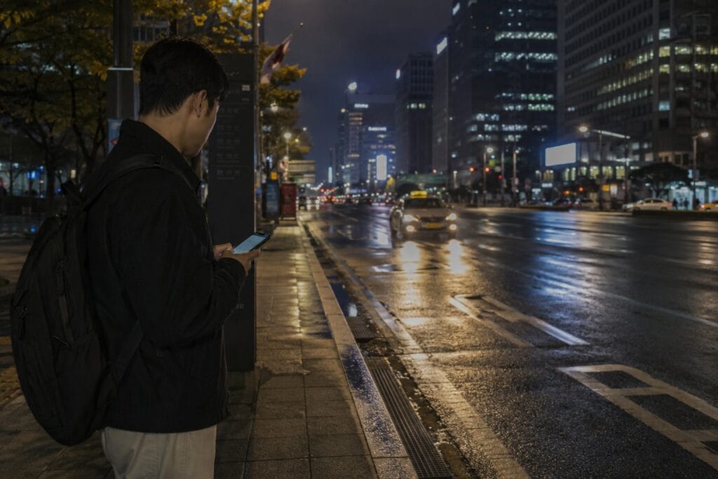 Traveler using a taxi app on a rainy night beside a main road in Seoul