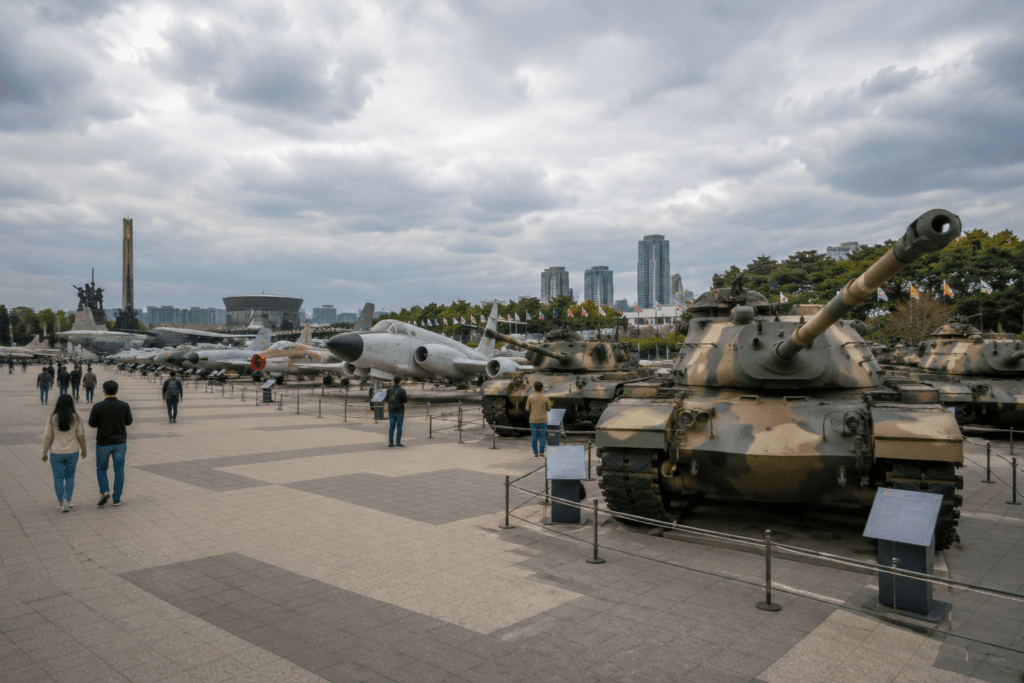 Outdoor military equipment display at a Seoul war memorial with tanks, aircraft, cloudy sky and visitors walking through the plaza